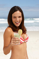 A young brunette woman on a beach with a smoothie wearing a bikini top