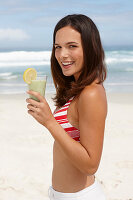 A young brunette woman on a beach with a smoothie wearing a bikini top and white trousers