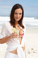 A young brunette woman on a beach with a smoothie wearing a white shirt and white trousers