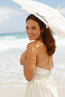 A brunette woman by the sea wearing a white summer dress and holding a parasol
