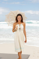 A brunette woman by the sea wearing a white summer dress and holding a parasol