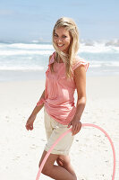 Junge Frau mit Hula-Hoop-Reifen im rosa Top und Shorts am Strand