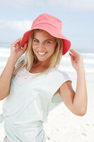 A blonde woman on the beach wearing a light t-shirt and a red hat