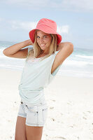 A blonde woman on the beach wearing a light t-shirt and denim shorts holding a red hat