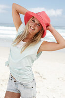 A blonde woman on the beach wearing a light t-shirt and a red hat