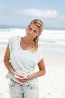 A blonde woman on the beach wearing a light t-shirt and holding a shell