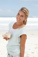 A blonde woman on the beach wearing a light t-shirt and holding a shell