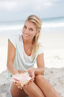 Blonde Frau mit Muschelschale in hellem T-Shirt am Strand