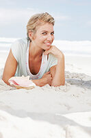 A blonde woman sitting on the beach wearing a light t-shirt with a shell in front of her