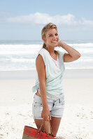 Blonde Frau mit Basttasche in hellem T-Shirt und Jeansshorts am Strand