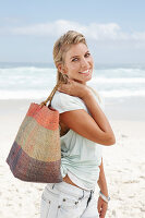 A blonde woman on the beach wearing a light t-shirt and holding a wicker bag