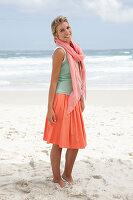 A blonde woman on a beach wearing a scarf, a turquoise top and a salmon-pink skirt