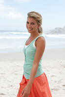 A blonde woman on a beach wearing a turquoise top and a salmon-coloured skirt