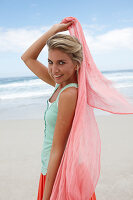 A blonde woman on a beach with a scarf wearing a turquoise top and a salmon-pink skirt