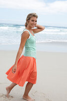 A blonde woman on a beach wearing a turquoise top and a salmon-coloured skirt