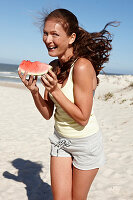Brünette Frau mit Wassermelone in hellem Top und Shorts am Strand