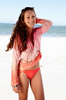 A brunette woman on a beach wearing a pink sequinned blouse and a bikini