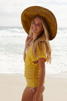 Blonde Frau mit Hut in gelbem Stricktop und Bikini am Strand