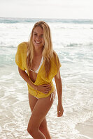 A blonde woman on a beach wearing a yellow knitted top and a bikini