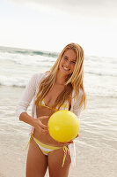 A blonde woman on a beach with a yellow ball wearing a white and yellow bikini and a cardigan