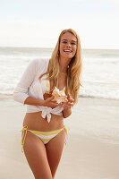 A blonde woman on a beach holding a shell wearing a white and yellow bikini and a cardigan