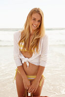 A blonde woman on a beach wearing a white and yellow bikini and a cardigan