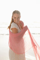 A blonde woman by the sea with a salmon-pink scarf and wearing a white dress