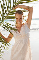 A blonde woman by the sea holding a palm frond wearing a white dress