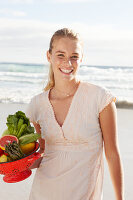 A blonde woman by the sea with a bowl of fruit and vegetables wearing a white dress