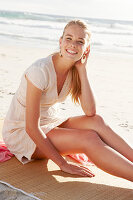 A blonde woman on a beach mat by the sea wearing a white dress