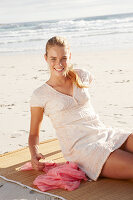 A blonde woman on a beach mat by the sea wearing a white dress