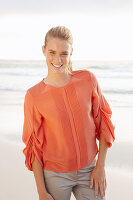 Junge blonde Frau in orangefarbener Bluse und heller Hose am Meer