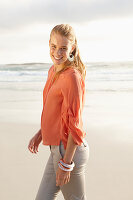 A young blonde woman wearing an orange blouse and light trousers by the sea