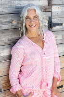 A mature woman wearing a pink blouse standing against a wooden wall