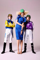 A blonde woman wearing a green hat and a blue dress with two men in jockey's outfits