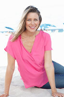 A blonde woman sitting in the sand wearing a pink blouse and jeans
