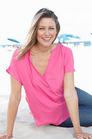 Blonde Frau in pinkfarbener Bluse und Jeans im Sand sitzend