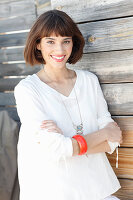 A brunette woman wearing a white long-sleeved blouse standing in front of a wooden wall