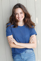 A young brunette woman in front of a grey wooden wall wearing a blue t-shirt