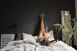 Young woman lying on double bed with legs stretched up dark grey wall