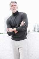 A grey-haired man on a terrace wearing a grey turtleneck jumper and light trousers