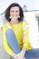 A young woman wearing a long-sleeved yellow top and jeans with a jumper over her shoulders