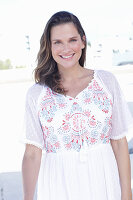 A long-haired woman wearing a white summer dress with colourful embroidery detail