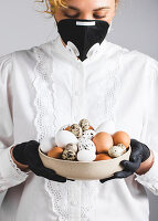A woman wearing a white blouse and a black face mask and gloves holding a bowl of eggs