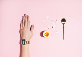 Frauenhand mit Armbanduhr, aufgebrochenes, gekochtes Ei und Eierlöffel