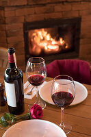 Zwei Rotweingläser mit Flasche und roter Rose auf Holztisch vor Kamin