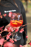 A hand holding a glass of Aperol Spritz with a sprig of blossom in the foreground