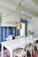 White dining table and chairs below pendant lamp and wood-burning stove against blue-striped wallpaper