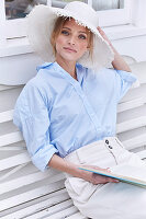 Blonde Frau mit Hut in hellblauer Hemdbluse und weißer Hose sitzt auf der Terrasse