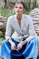 A blonde woman wearing a white blazer and a denim skirt sitting on a fallen tree